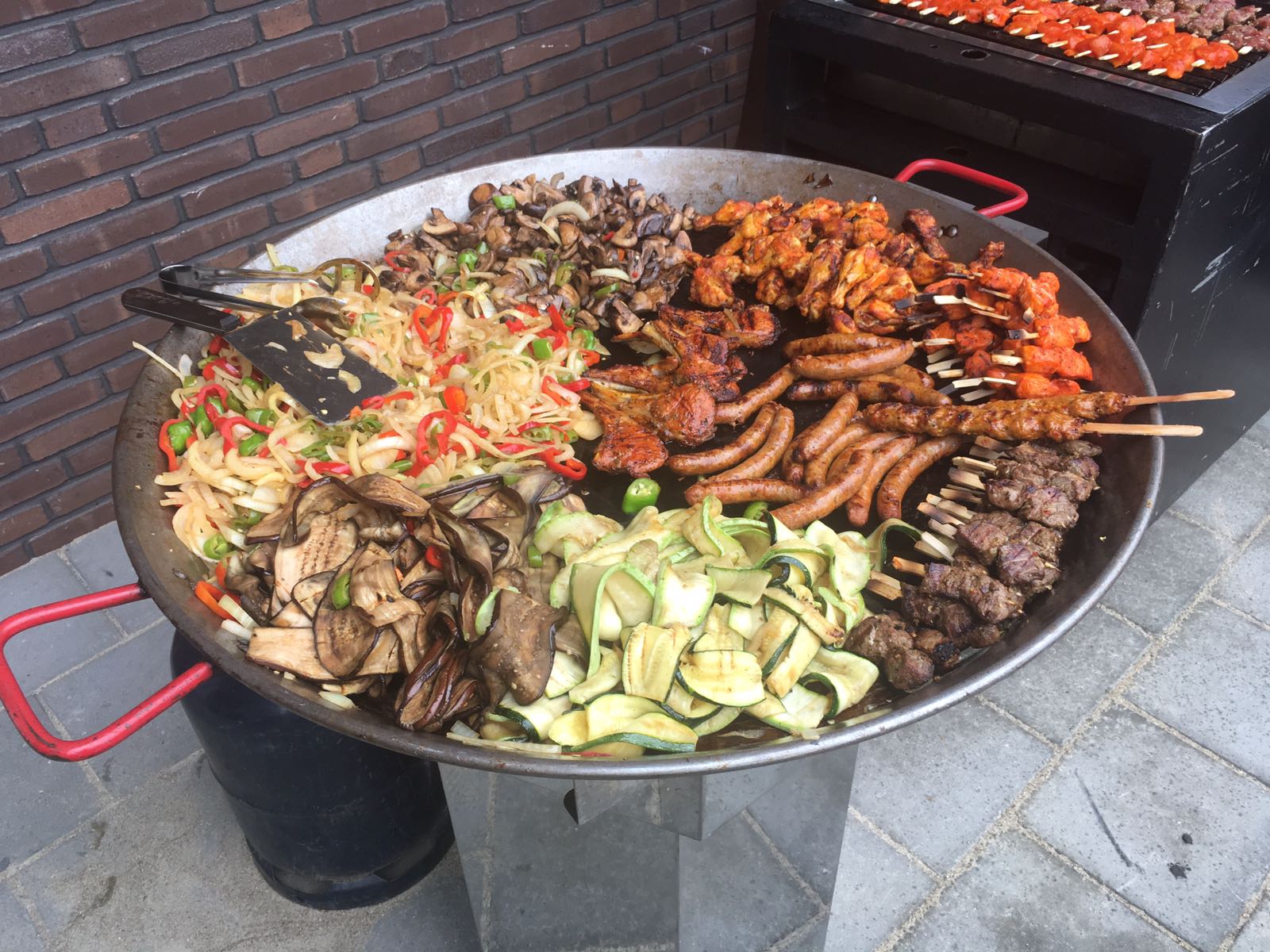 Een grote BBQ-pan buiten de winkel met gegrilde groenten, worst en spiesen
